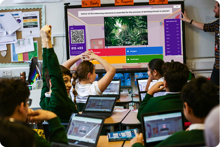 Primary school students raising hands and using laptops during a Kahoot science quiz on a classroom smartboard 
